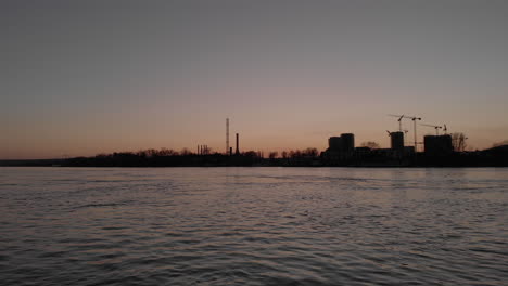 Push-in-drone-flying-video-over-a-river-as-cranes-constructing-industrial-buildings,-nuclear-power-plants-at-sunset