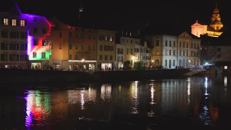Static-wide-shot-of-illuminated-restaurants-at-riverbank-during-night-with-church-in-background
