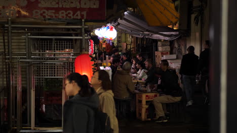 Tracking,-slow-motion-shot-of-people-sitting-at-a-restaurant,-while-others-walk-on-the-Ueno-shopping-street,-at-night-time,-in-Tokyo,-Japan