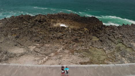 Aerial-view-of-caucasian-couple-at-the-coast-watching-the-beautiful-tropical-blue-seascape-with-waves-crashing-into-the-rocks-in-Bali