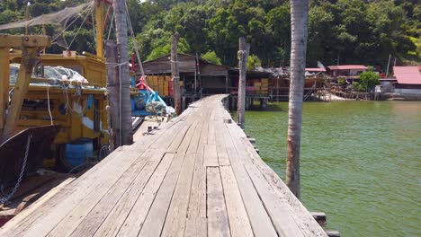 A-narrow-wooden-bridge-on-the-water-with-ships,-houses-and-green-forest-at-the-background-in-Pangkor-Island-Malaysia