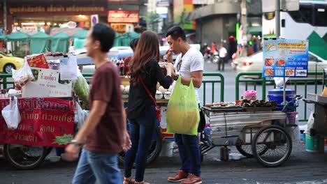 Ein-Mann-Fotografiert-Eine-Frau-Auf-Dem-Bürgersteig-Von-Bangkok,-Thailand,-Mit-Straßenverkäufern-Und-Vorbeifahrenden-Autos-Im-Hintergrund-–-Totale