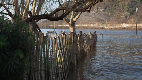 Alter-Holzzaun,-Bäume-Und-Straßenlaternen-Stehen-Im-Hochwasser-Mit-Fluss-Im-Hintergrund