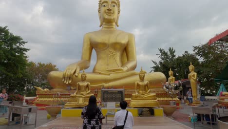 Toma-Inclinada-De-Una-Estatua-Dorada-Del-Gran-Buda-En-Pattaya-Con-Algunas-Personas-Rezando-Frente-Al-Monumento-Sagrado.