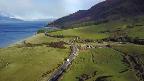 Aerial-video-of-a-Van-driving-on-a-Coastal-road-with-green-paddocks,-then-panning-to-the-blue-ocean,-and-Snowy-mountains