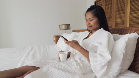 Adult-woman-drinking-coffee-and-using-her-mobile-phone,-browsin-the-internet-while-laying-on-a-white-bed-wearing-a-white-dressing-gown