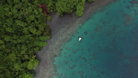 Eine-Sich-Drehende-Drohnenaufnahme-Eines-Kleinen-Weißen-Bootes,-Das-Am-Ufer-Einer-Kleinen-Insel-Mit-Grauen-Felsen,-üppigen-Grünen-Bäumen-Und-Einem-Farbenfrohen-Korallenriff-Darunter-Ankert
