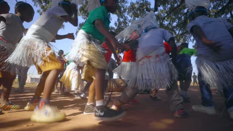 Group-of-young-African-local-school-children-are-dancing-a-traditional-dance-with-costumes-in-a-circling-while-stepping-on-the-rhythm-beat-of-the-drum-in-dirt-and-rural-bush-nature-of-Ghana-slowmotion