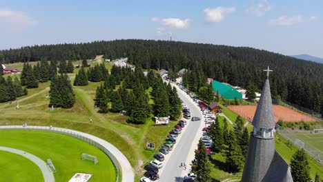 The-Podruznicna-church-with-vehicles-filling-the-street-for-an-event-near-the-soccer-field,-Aerial-flyover-shot