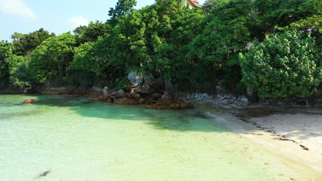 Exotic-beach-under-shadow-of-tropical-trees-and-lush-vegetation,-clear-calm-water-of-lagoon-washing-limestone-cliffs,-Thailand