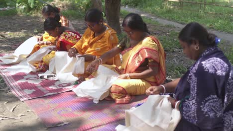 Rural-Indian-tribal-women-sewing-cloths-with-needles-and-thread-sitting-on-ground-outdoor,-feminism-and-self-dependent,-non-profit-organization