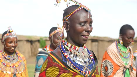 Stammes-Maasai-Frauen,-Die-Tanz-Und-Gesang-In-Nahaufnahme-Aufführen,-Mit-All-Ihrem-Traditionellen-Schmuck-Im-Hintergrund-Sind-Die-Lehmhäuser,-In-Denen-Sie-Im-Masai-Mara-Nationalpark-In-Kenia,-Afrika,-Leben
