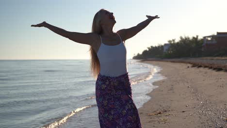 Facing-the-camera-a-mature-backlit-woman-raises-her-arms-and-looks-to-the-sky-while-on-a-beach