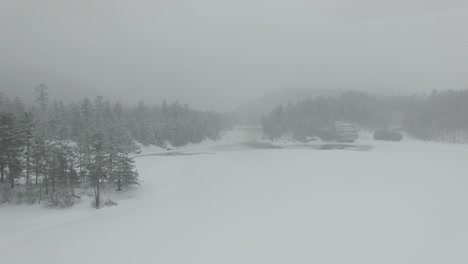Drone-flying-over-lake-covered-in-white-ice-during-snow-storm-towards-forest