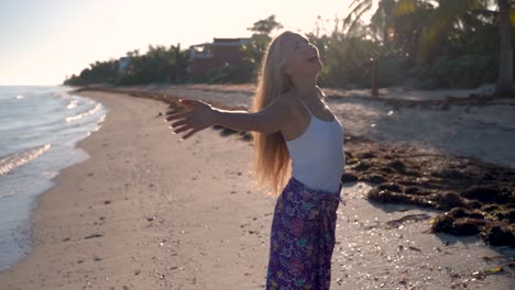 Mature-blonde-woman-raises-her-arms-and-looks-to-the-sky-while-on-a-beach-as-the-camera-turns-around-her