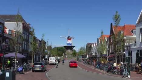 Historical-windmill-wrapped-in-Dutch-flag,-situated-at-the-end-of-a-shopping-street-full-of-parked-bikes-and-historical-houses-in-the-Netherlands-on-a-sunny-summer-day