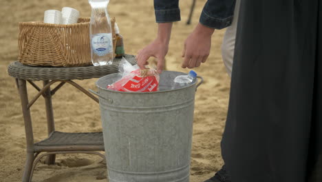 Person-Empty’s-Bag-Of-Ice-Into-Metal-Bucket-On-The-Beach,-SLOW-MOTION