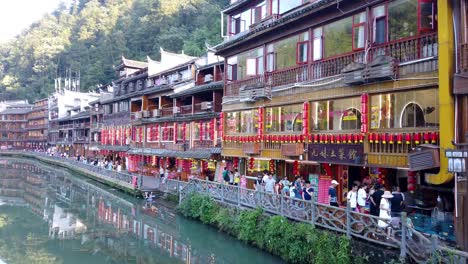 people-and-tourists-walking-in-front-of-traditional-old-wooden-houses-on-the-riverbanks-of-Tuo-river,-flowing-through-the-centre-of-Feng-huang-Old-Town