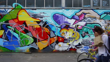 Mit-Graffiti-Bedeckte-Wand-Im-Rohen-Gelände,-Vorbeigehende-Menschen