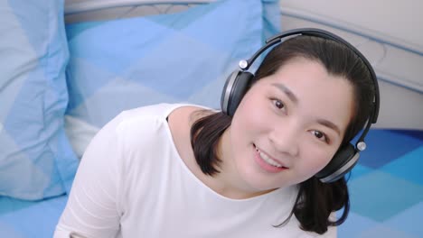 Mujer-Asiática-Sonriendo-Usando-Los-Auriculares-Bluetooth-Para-Escuchar-Música-Y-Mirando-La-Cámara-Tan-Encantadora-Tumbada-En-La-Cama