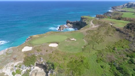 Scenic-view-of-rocky-seashore-landscape,-green-golf-course-greens-above-edge-of-cliffs-and-sandy-pits-by-blue-ocean-water,-Macao,-China,-overhead-aerial-pull-back