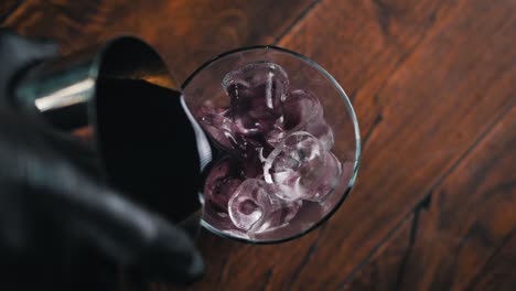 Bartender-wearing-black-gloves-is-pouring-a-purple-drink-into-a-glass-filled-with-ice-cubes