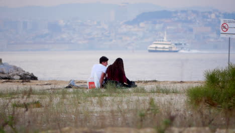 Young-Adult-Couple-Sitting-On-Beach-Sand-With-A-Passenger-Boat-Sailing-In-Background