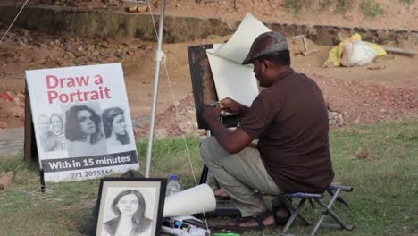 Street-Sketch-Artist-getting-ready-for-pencil-Portraiture-drawing-of-a-small-child-on-a-piece-of-white-paper,-sitting-on-a-small-foldable-chair-under-a-big-umbrella,-Galle-Dutch-Fort-b-roll-clip