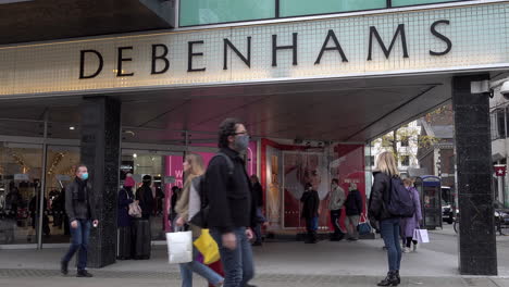 A-left-to-right-camera-truck-technique-passes-a-tree-trunk-and-reveals-people-entering-and-exiting-Debenhams-department-store-on-Oxford-Street-as-others-walk-by-carrying-shopping-bags