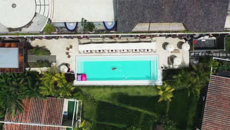 Woman-in-red-swimsuit-swimming-at-pool-on-Shore-Amora-Canggu-Hotel-pool-in-Bali,-Indonesia,-Aerial-rising-full-reveal-shot