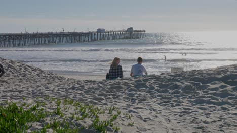 Couple-sitting-on-the-beach,-next-to-a-pier,-in-San-Clemente,-California