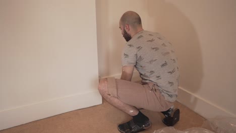 Young-caucasian-adult-man-on-his-knees-painting-white-wall-of-empty-room-in-house-with-roller-cambridge-england