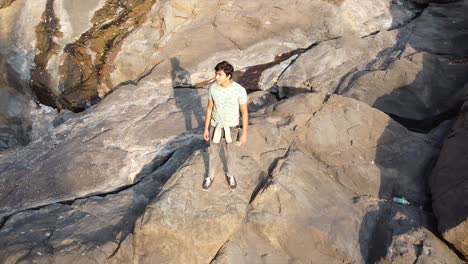 Aerial-view-or-drone-shot-of-a-young-Asian-boy-standing-on-a-cliff-of-small-mountain-made-with-rocks-in-forest-near-a-small-waterfall-video-background