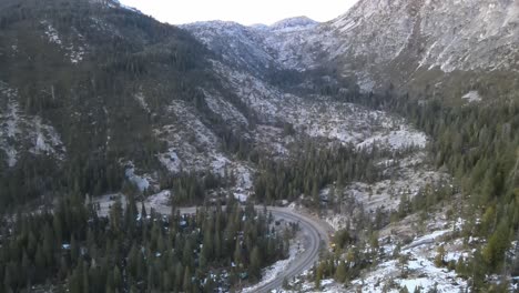 Flying-on-a-winter-day-nearby-a-asphalt-road-in-a-valley-shaded-by-the-mountains-with-pine-trees-and-snow-on-the-ground