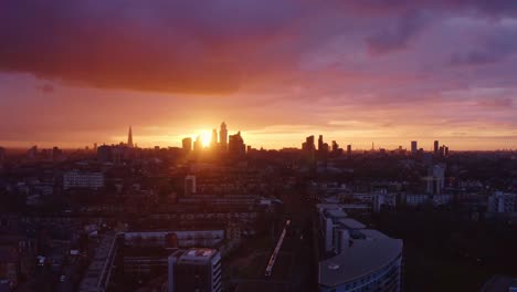 Aerial-drone-shot-towards-city-of-London-skyscrapers-as-train-passes-underneath-sunset