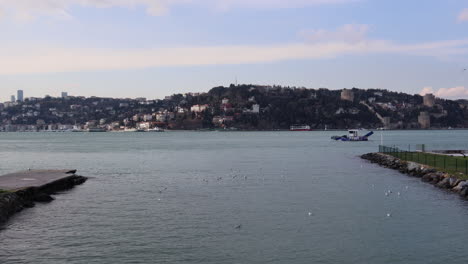 Möwen-Schwimmen-Am-Bosporus-Mit-Einem-Segelboot---Rumelihisari,-Rumelische-Burg-Mit-Üsküdar-Am-Ufer-Des-Bosporus-In-Istanbul,-Türkei