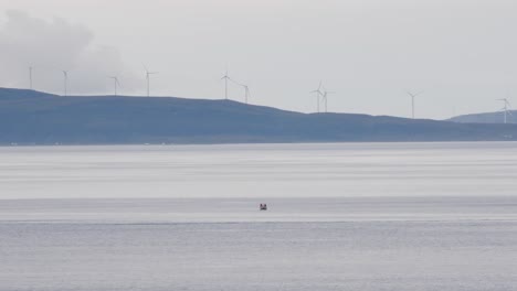 Static-wide-shot-of-fisherman-in-fishing-boat-on-calm-ocean-and-hills-landscape-with-rotating-windmills-in-background
