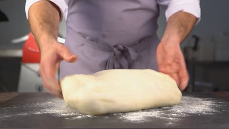 Ein-Bäcker-Bei-Der-Arbeit,-Der-Eine-Graue-Schürze-Trägt-Und-Mit-Seinen-Händen-Einen-Großen-Teig-Knetet,-Um-Brot,-Focaccia-Oder-Pizza-Zuzubereiten