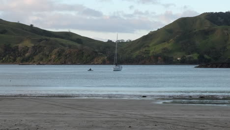 Two-people-paddling-on-tender-to-reach-sail-boat-on-bay,-new-zealand
