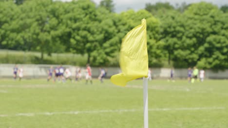 Una-Bandera-De-Esquina-Amarilla-Ondeando-En-El-Viento-En-Un-Campo-De-Fútbol,-Equipos-De-Fútbol-Femenino-Jugando
