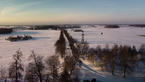 Beautiful-aerial-birdseye-view-of-snow-covered-fields-and-rural-gravel-road-in-sunny-winter-day,-golden-hour,-car-on-the-road,-wide-angle-drone-shot-moving-forward