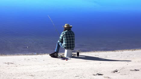 Un-Pescador-Solitario-Sentado-En-Una-Playa-Enrolla-Lentamente-Su-Línea-Después-De-Perder-Su-Cebo