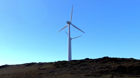 Two-wind-turbines-with-their-blades-spinning-at-the-top-of-the-mountain-on-a-quiet,-sunny-afternoon-with-clear-blue-skies