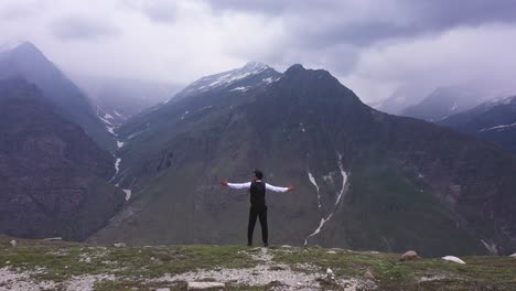 Aerial-shot-of-man-standing-in-the-edge-of-one-mountain-with-a-surrounded-mountain-in-the-background