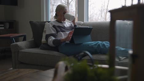 Woman-sitting-on-the-couch-in-front-of-a-bay-window,-drinking-coffee-while-scrolling-on-a-tablet-with-her-feet-up-on-the-couch-from-a-diagonal-angle
