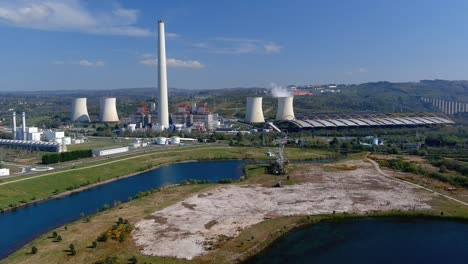 Thermal-power-plant-with-smoking-chimney-behind-the-lake-and-an-old-drill,-bridge-on-the-right,-bright-and-sunny-afternoon