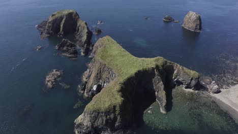 Aerial-descends-to-reveal-eroded-sea-caves-in-jagged-islet-in-blue-sea