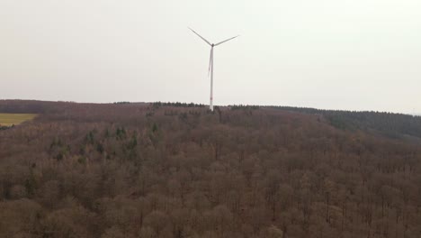 Aerial-footage-of-approaching-flight-towards-a-fast-spinning-wind-turbine-towering-on-top-of-a-brown-hill-covered-by-leafless-deciduous-trees