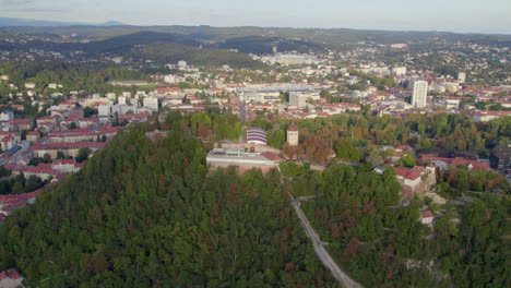 Aerial-view-above-landmark-Glokenturm-tower-on-Graz's-SchloÃŸberg-hilltop-woodland-park-panning-across-cityscape