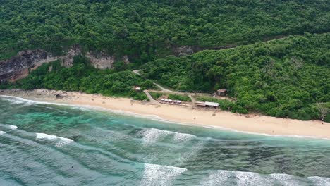small-restaurant-on-empty-white-sand-beach-of-Nyang-Nyang-in-Uluwatu-Bali-with-ocean-waves-crashing,-aerial